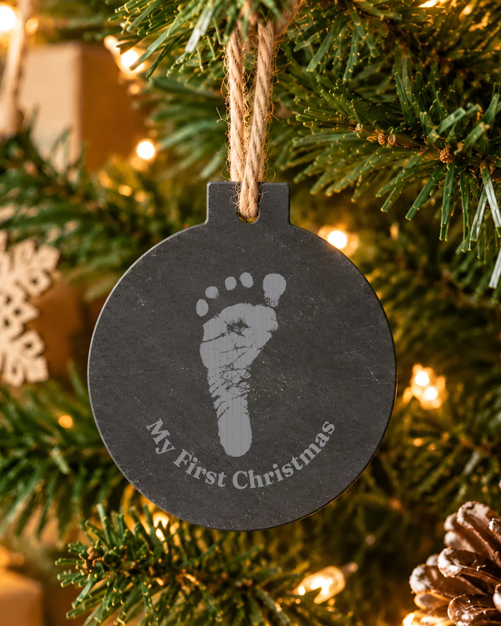 A natural slate Baby's First Christmas bauble hanging on a Christmas tree, engraved with a baby's footprint and My First Christmas